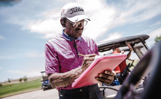 Driver of single seater golf buggies stands in white cap and pink stripey shirt marking off purchases on a clipboard.